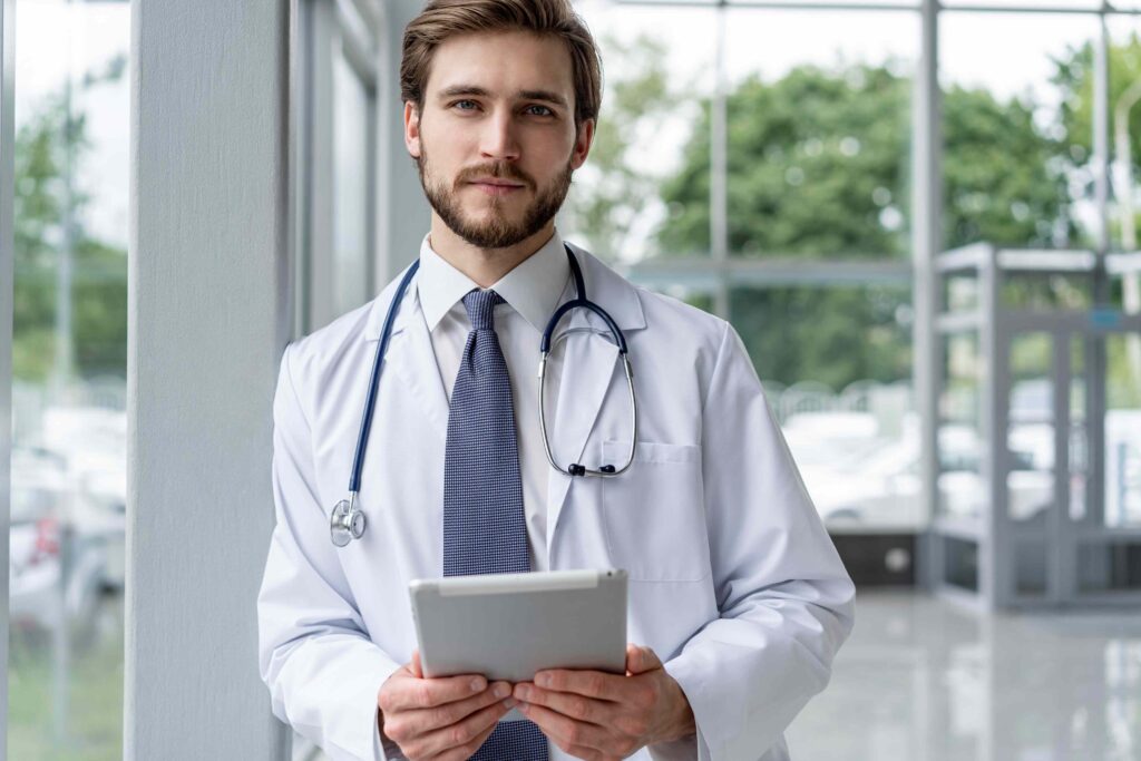 Arzt in weißem Kittel mit Stethoskop und Tablet vor moderner Klinik.